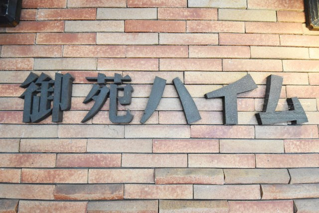 【現況写真】 | 御苑ハイム | エントランスに掲げられた、マンション名を示す館銘板。さほど激しく主張はしませんが、しっかりと掲示されています。～即日のご見学可能～　お問合せはお気軽にどうぞ。
